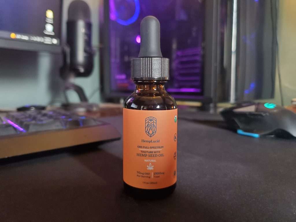 A bottle of HempLucid Full-Spectrum CBD oil with hemp seed oil, placed on a desk with a computer monitor, microphone, and gaming mouse in the background.