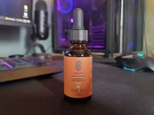 A bottle of HempLucid Full-Spectrum CBD oil with hemp seed oil, placed on a desk with a computer monitor, microphone, and gaming mouse in the background.